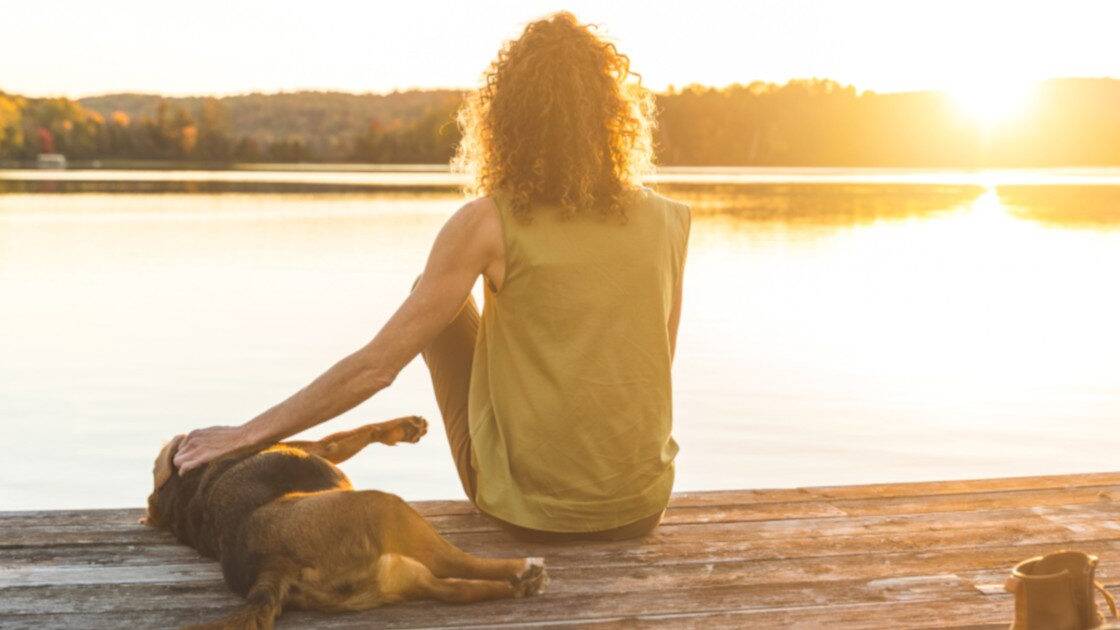 Anxiety and Depression - Lady with her dog sitting by a lake at sunset, the dog laying down next to her with her hand on its back, sharing a peaceful moment.