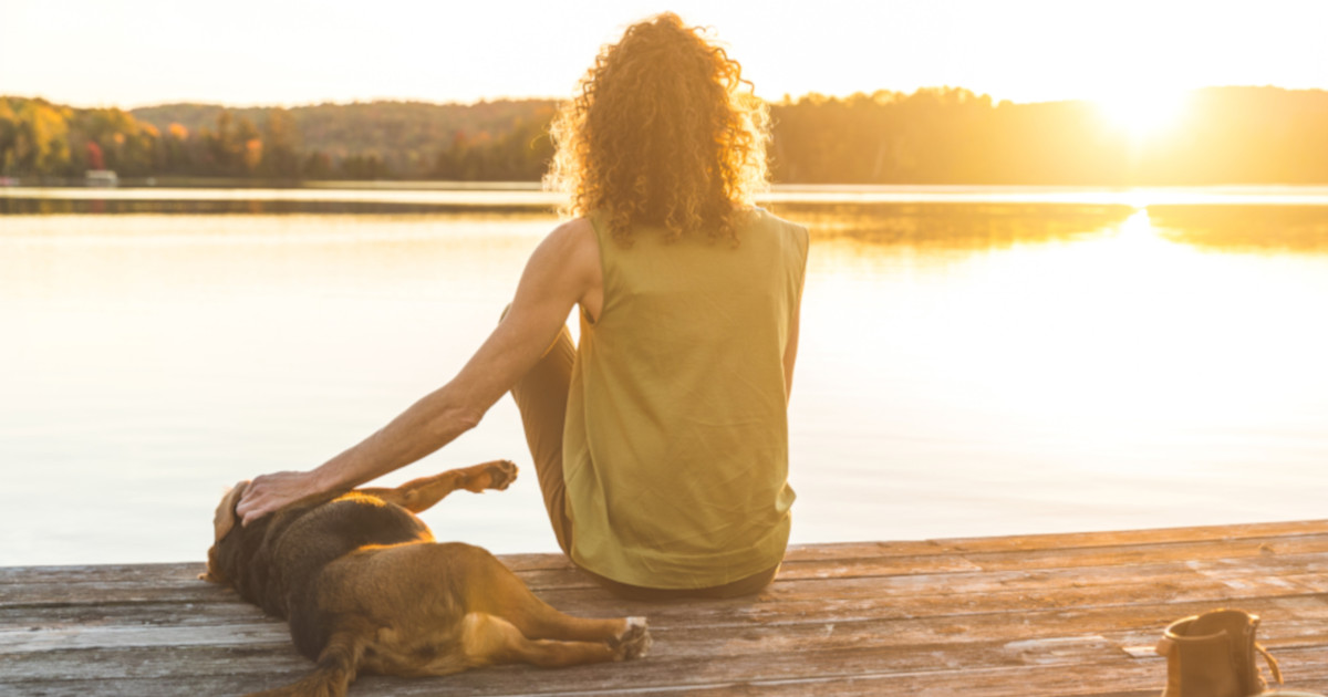 Anxiety and Depression - Lady with her dog sitting by a lake at sunset, the dog laying down next to her with her hand on its back, sharing a peaceful moment.