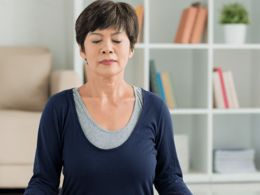 Stress Management - Middle-aged Asian woman sitting in meditation pose in her living room, practicing mindfulness exercises for blood pressure management.