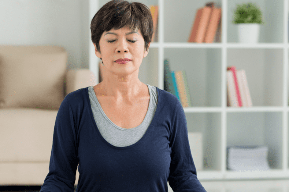 Stress Management - Middle-aged Asian woman sitting in meditation pose in her living room, practicing mindfulness exercises for blood pressure management.