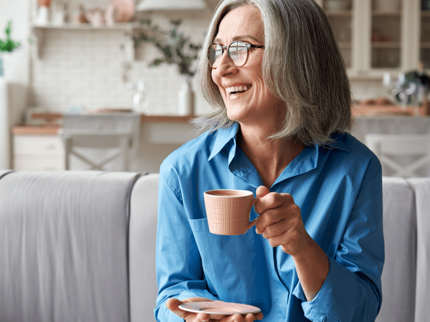 Senior woman enjoying her morning coffee on a couch, bathed in natural sunlight, symbolizing a relaxed approach to monitoring blood pressure.
