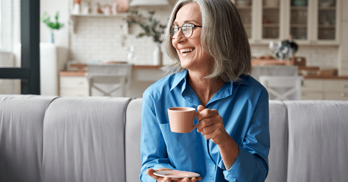 Senior woman enjoying her morning coffee on a couch, bathed in natural sunlight, symbolizing a relaxed approach to monitoring blood pressure.