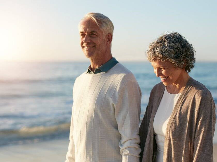 Happy senior couple, a lady and her husband in their 50s and 60s, walking hand-in-hand on the beach at sunrise, embodying vitality and heart health.