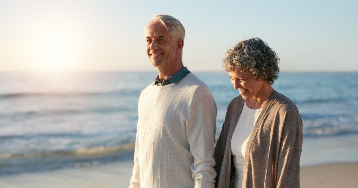 Happy senior couple, a lady and her husband in their 50s and 60s, walking hand-in-hand on the beach at sunrise, embodying vitality and heart health.