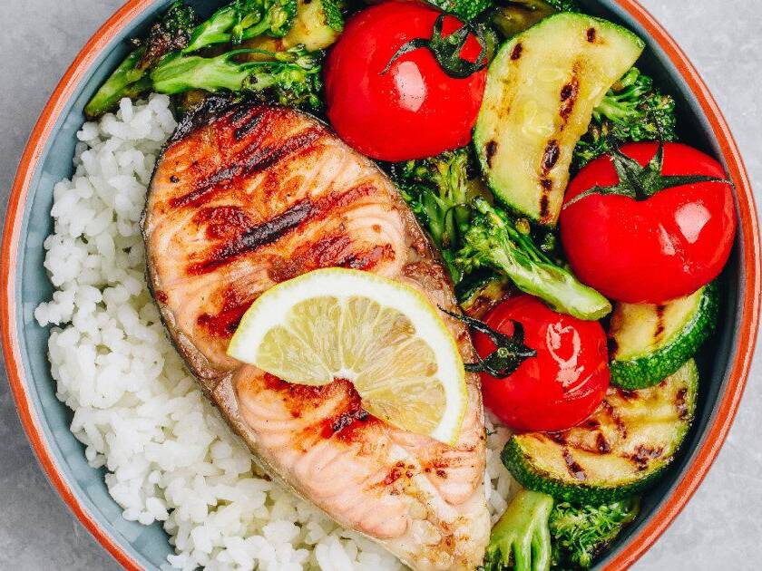 A nutritious meal showcasing the Mediterranean diet with white rice, grilled salmon, slices of avocado, tomato, and cucumbers on a plate.