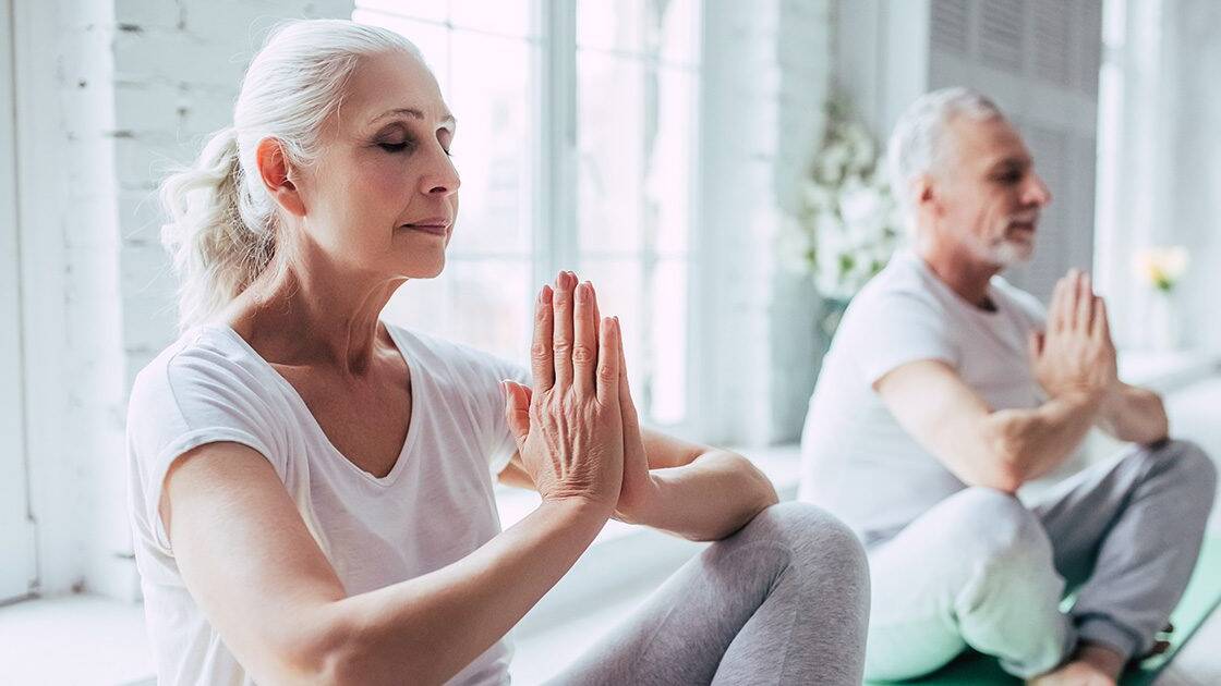 Mindfulness and Heart Health: A senior woman and man practicing yoga and meditation indoors with sunlight streaming through the window.