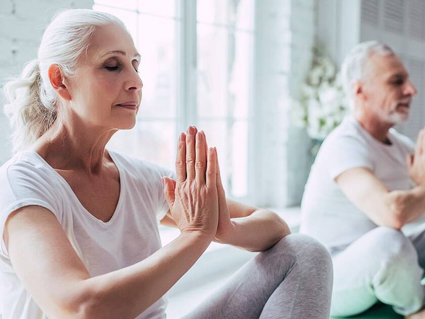 Mindfulness and Heart Health: A senior woman and man practicing yoga and meditation indoors with sunlight streaming through the window.