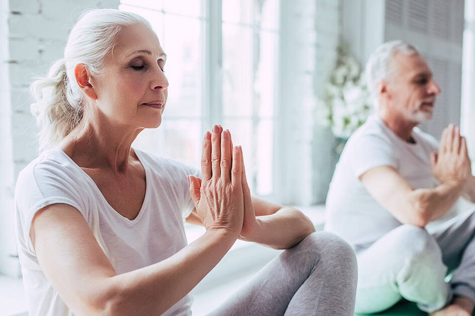 Mindfulness and Heart Health: A senior woman and man practicing yoga and meditation indoors with sunlight streaming through the window.