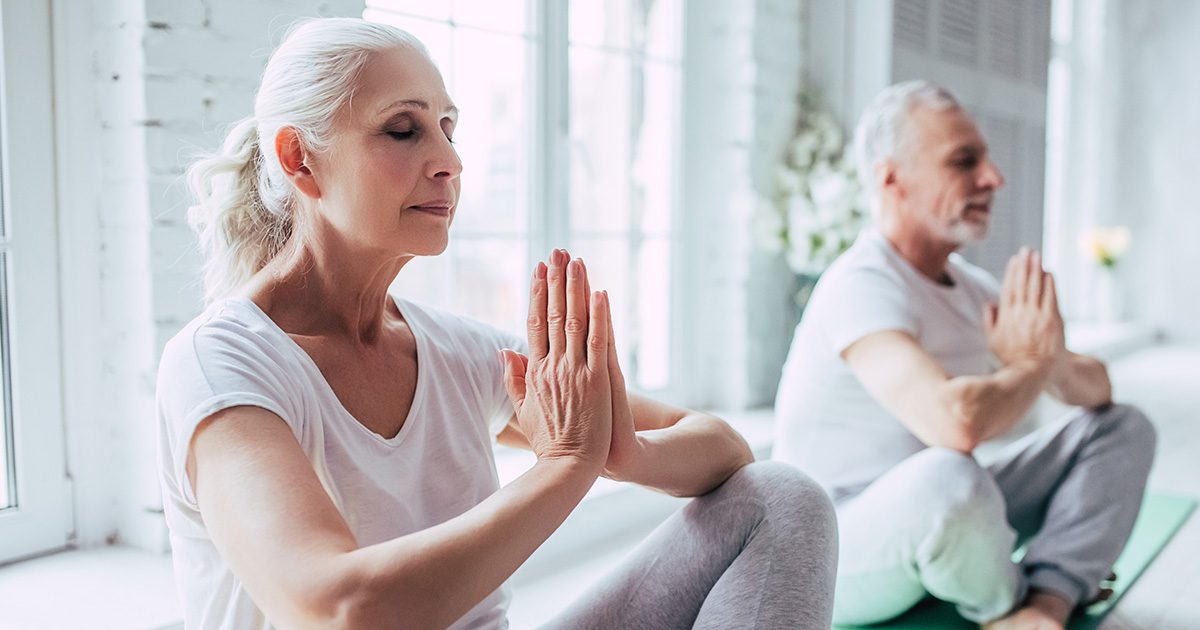 Mindfulness and Heart Health: A senior woman and man practicing yoga and meditation indoors with sunlight streaming through the window.