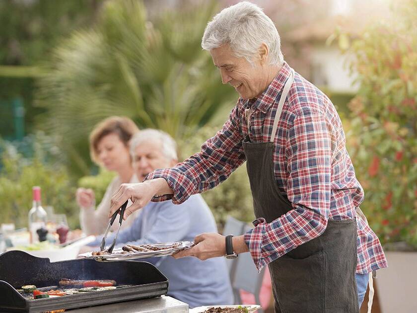 Community and Heart Health: Happy senior man grilling food on a barbecue with a woman sitting in the background, preparing to eat as a group outdoors during the day.