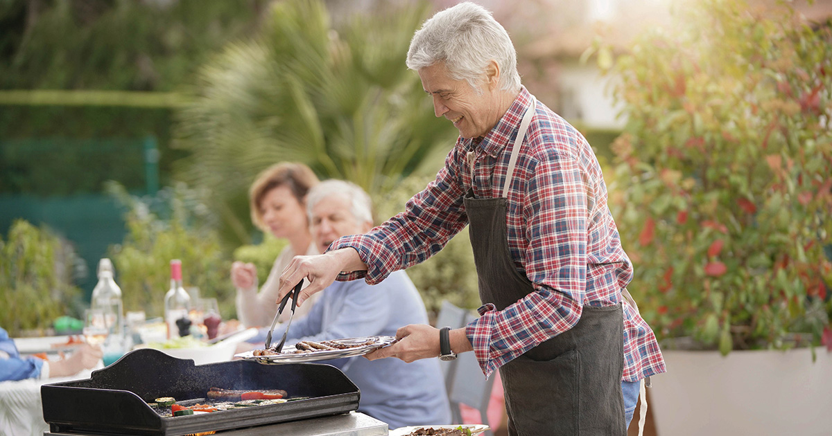 Community and Heart Health: Happy senior man grilling food on a barbecue with a woman sitting in the background, preparing to eat as a group outdoors during the day.